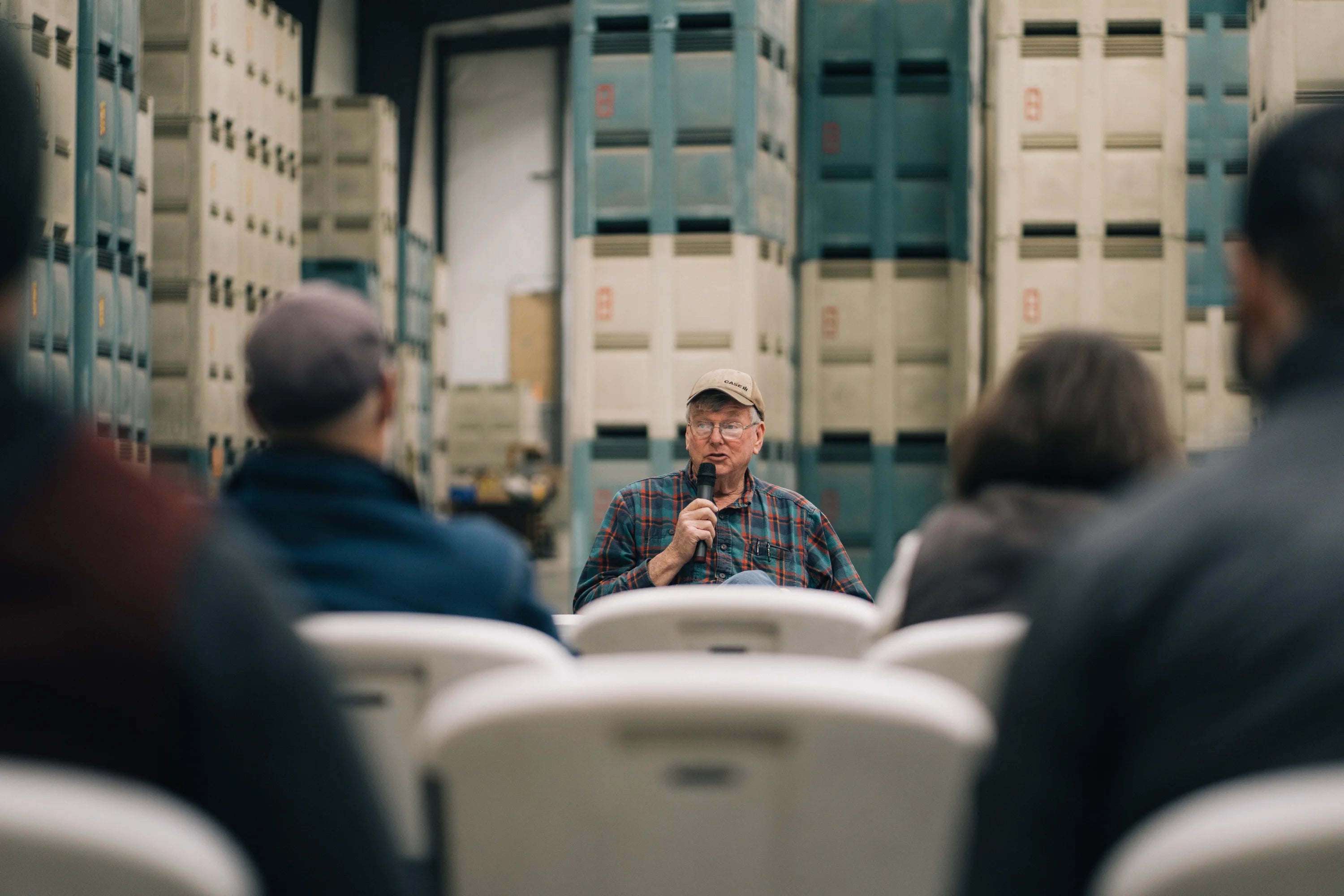 Person speaking into a microphone in a warehouse setting with stacked crates in the background.